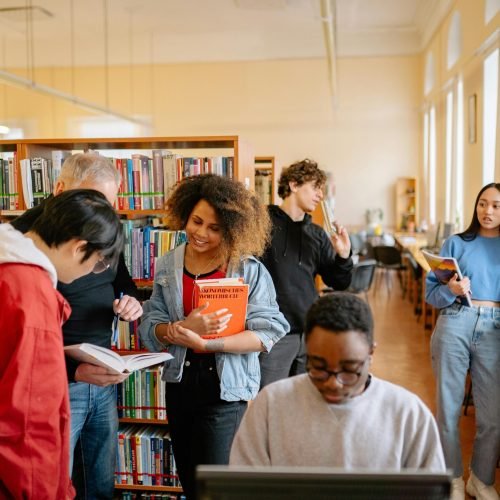 A diverse group of students studying together at a university library, engaging with books and technology.